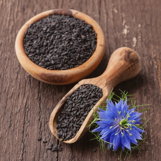 Wooden bowl and scoop filled with black seeds on a wooden surface with a blue nigella sativa flower.